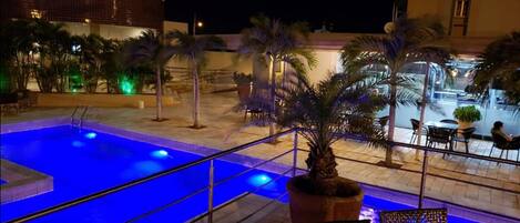 A nighttime view of a pool area with a blue-lit pool, palm trees, and outdoor seating.