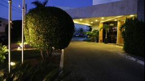 A paved pathway leading to a building entrance with a well-lit entrance area, surrounded by greenery and palm trees. - Garbos Trade Hotel (Mossoro)