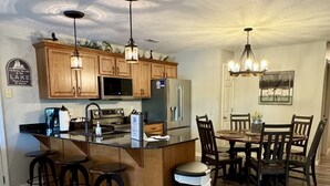 A kitchen with wooden cabinets, a stainless steel refrigerator, a dining table with chairs, and a trash can.