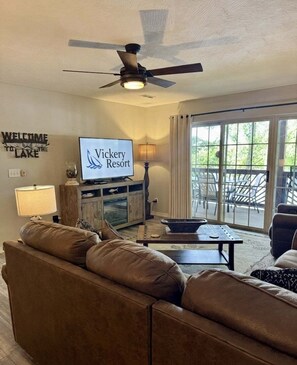 A living room with a sofa, a television displaying 'Vickery Resort,' a ceiling fan, and a view through large sliding glass doors.