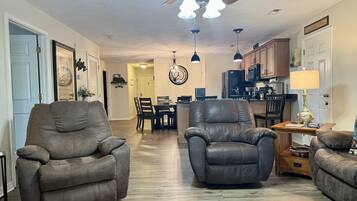 A spacious living room with two leather armchairs, a dining area with a wooden table and chairs, a kitchen with wooden cabinets, and a ceiling fan.