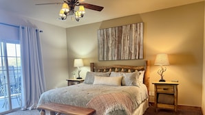 Une chambre avec un grand lit, un banc en bois, deux tables de chevet avec des lampes, un ventilateur de plafond et une grande fenêtre avec des rideaux.