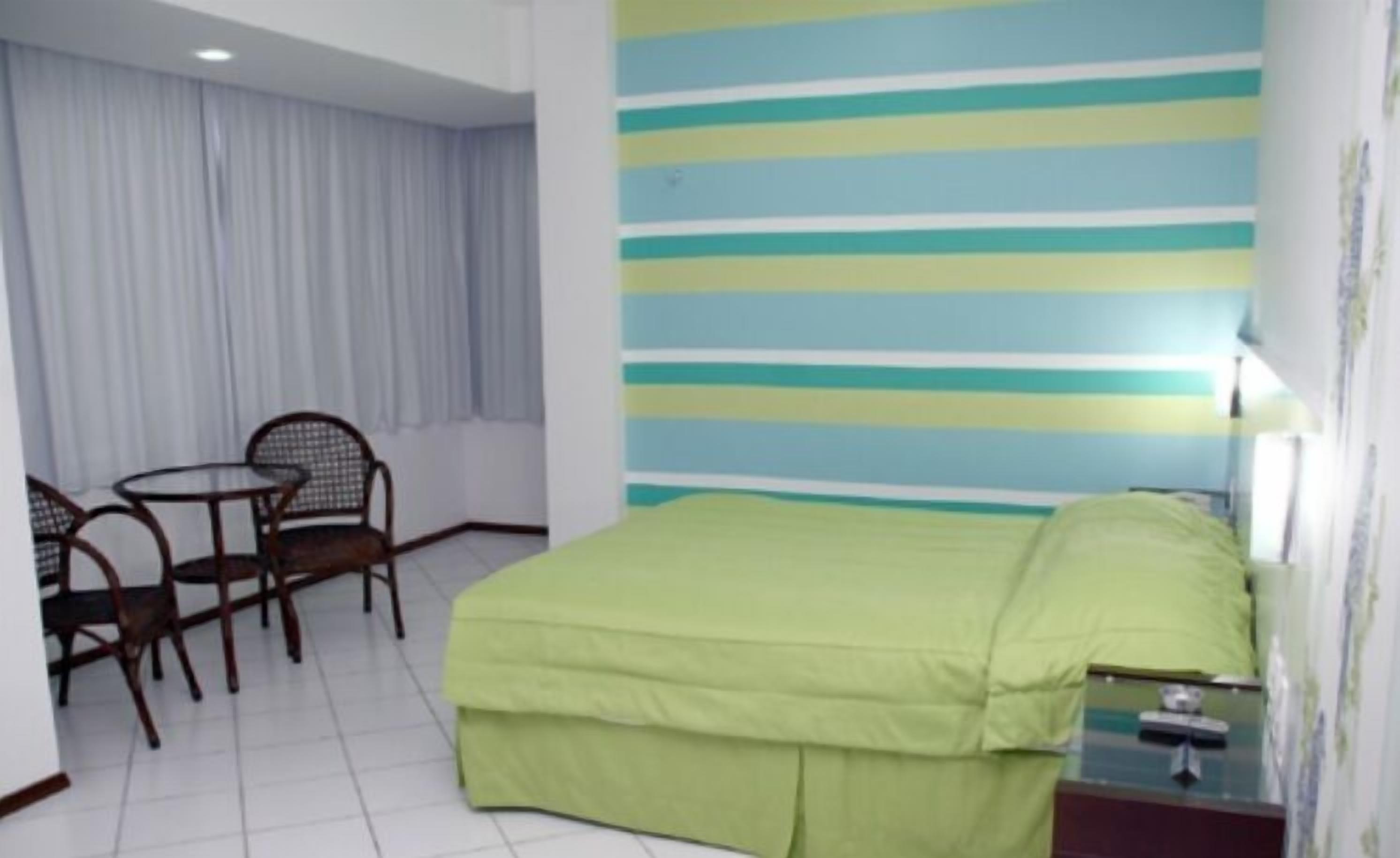 Une chambre d’hôtel avec un lit, une petite table, deux chaises, une fenêtre avec des rideaux et un mur à rayures.