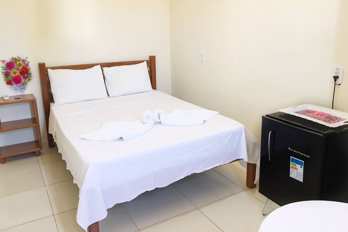Una habitación de hotel con una cama, una mesita de noche, un refrigerador pequeño y un florero con flores.
