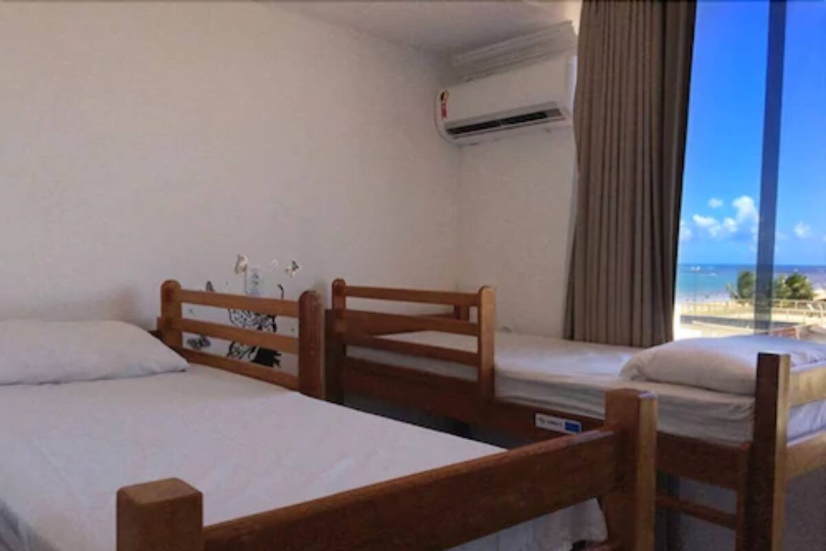 Una habitación con dos camas de madera, un ventanal con vista a la playa y un equipo de aire acondicionado en la pared.