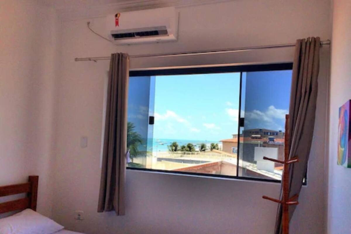 Habitación de hotel con cama, ventana con cortinas, equipo de aire acondicionado y vista a edificios y palmeras.