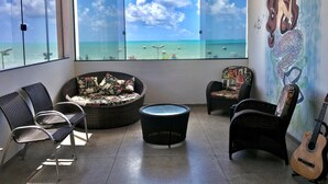 A room with a large window overlooking a beach, wicker furniture, and a guitar. - Maraga Beach Hostel (Maragogi)