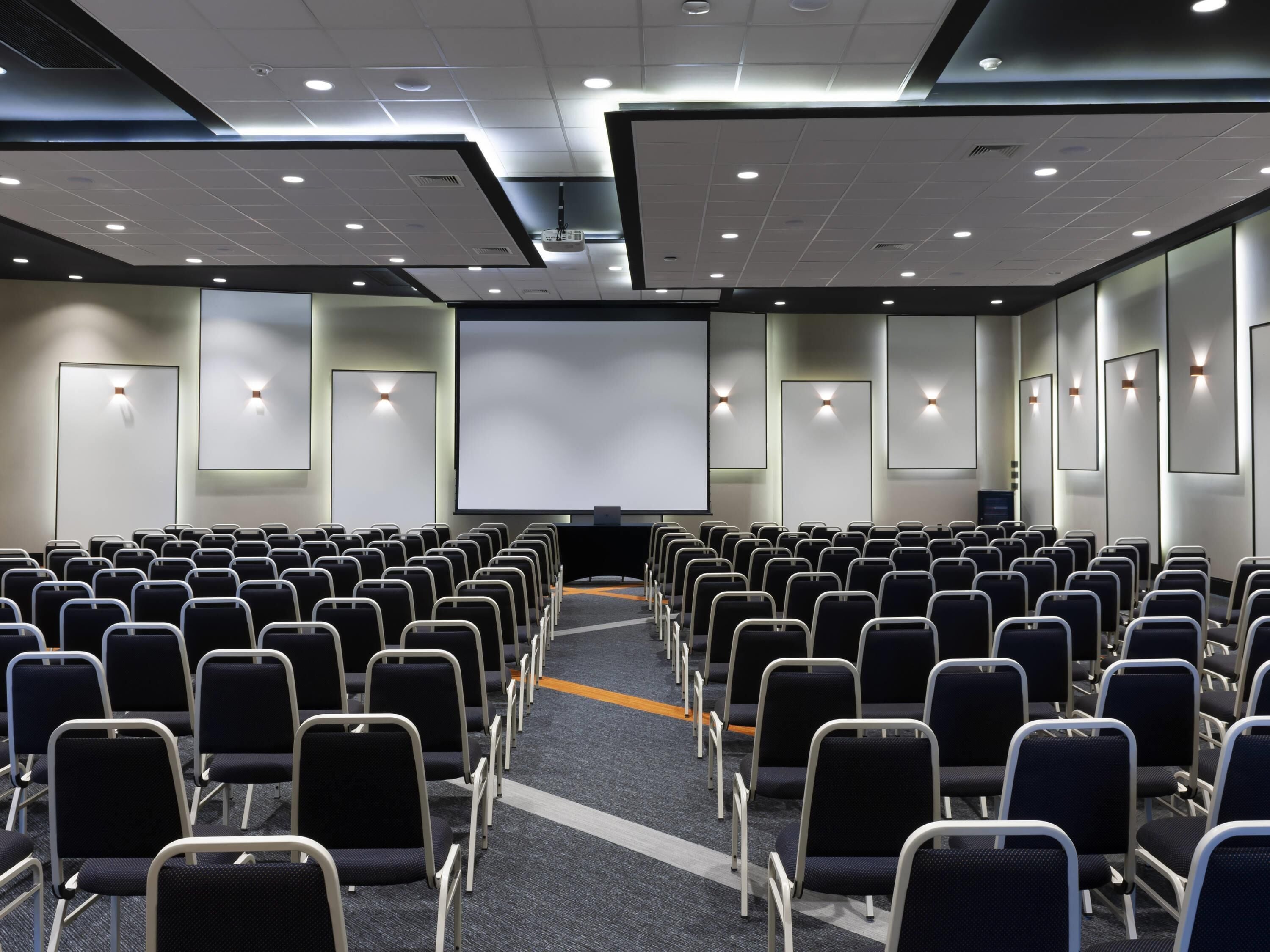 Un salón de conferencias con filas de sillas, una pantalla grande y un proyector.