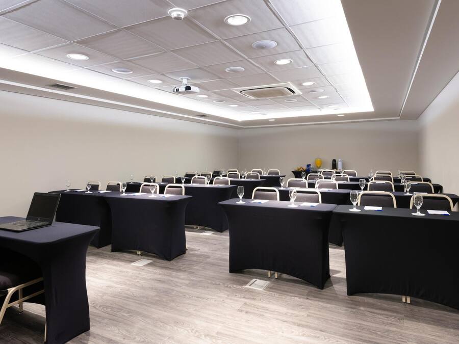 Sala de conferência com fileiras de mesas, cadeiras, um projetor e iluminação embutida no teto.