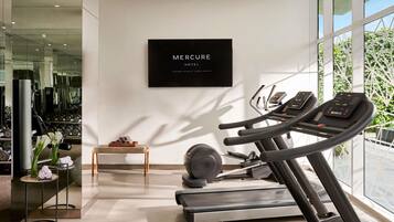 A modern gym with treadmills, a wall-mounted TV displaying 'Mercure Hotel Jakarta PANTAI INDAH KAPUK,' and a large window with greenery outside.