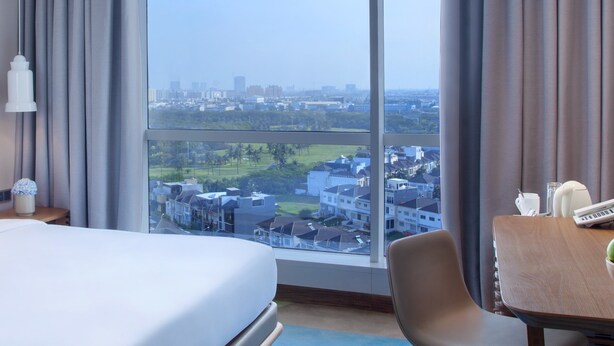 Une chambre d’hôtel avec un grand lit, un bureau avec une chaise et une vue sur le paysage urbain grâce à de grandes fenêtres.