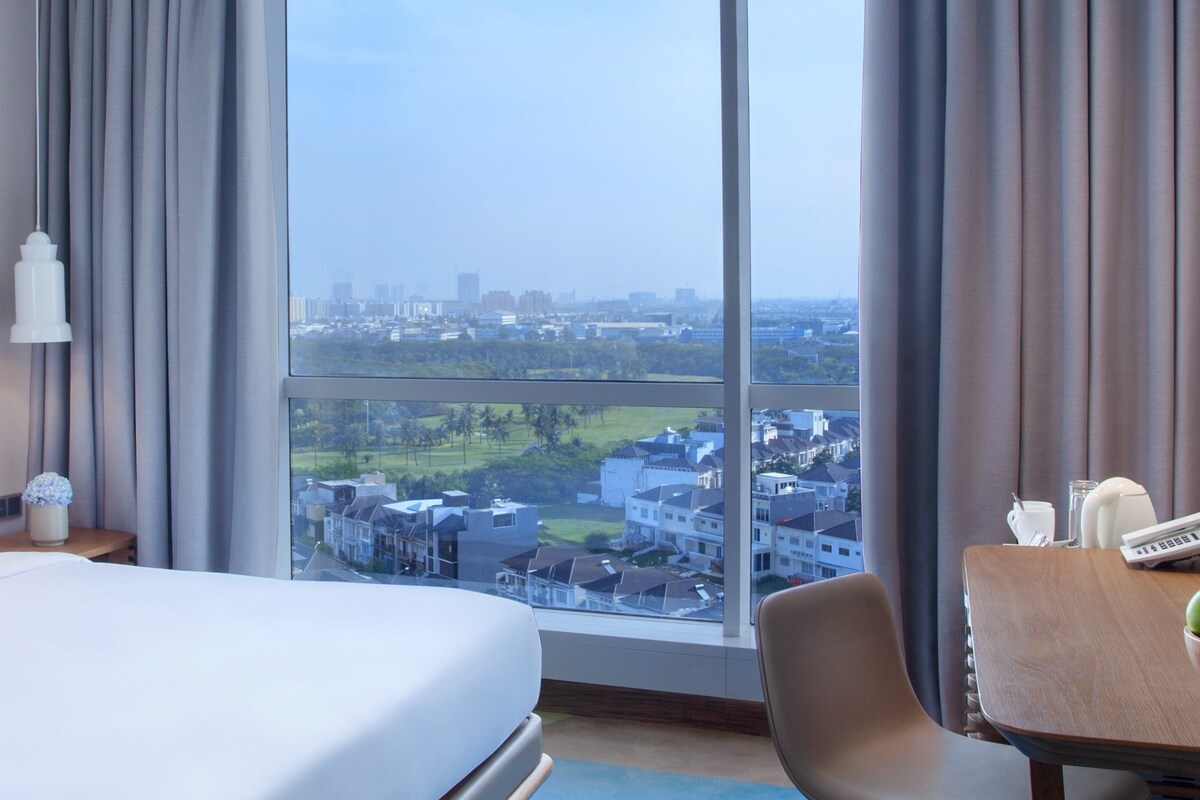 Habitación de hotel con una cama grande, un escritorio con silla y vistas a un paisaje urbano a través de amplias ventanas.