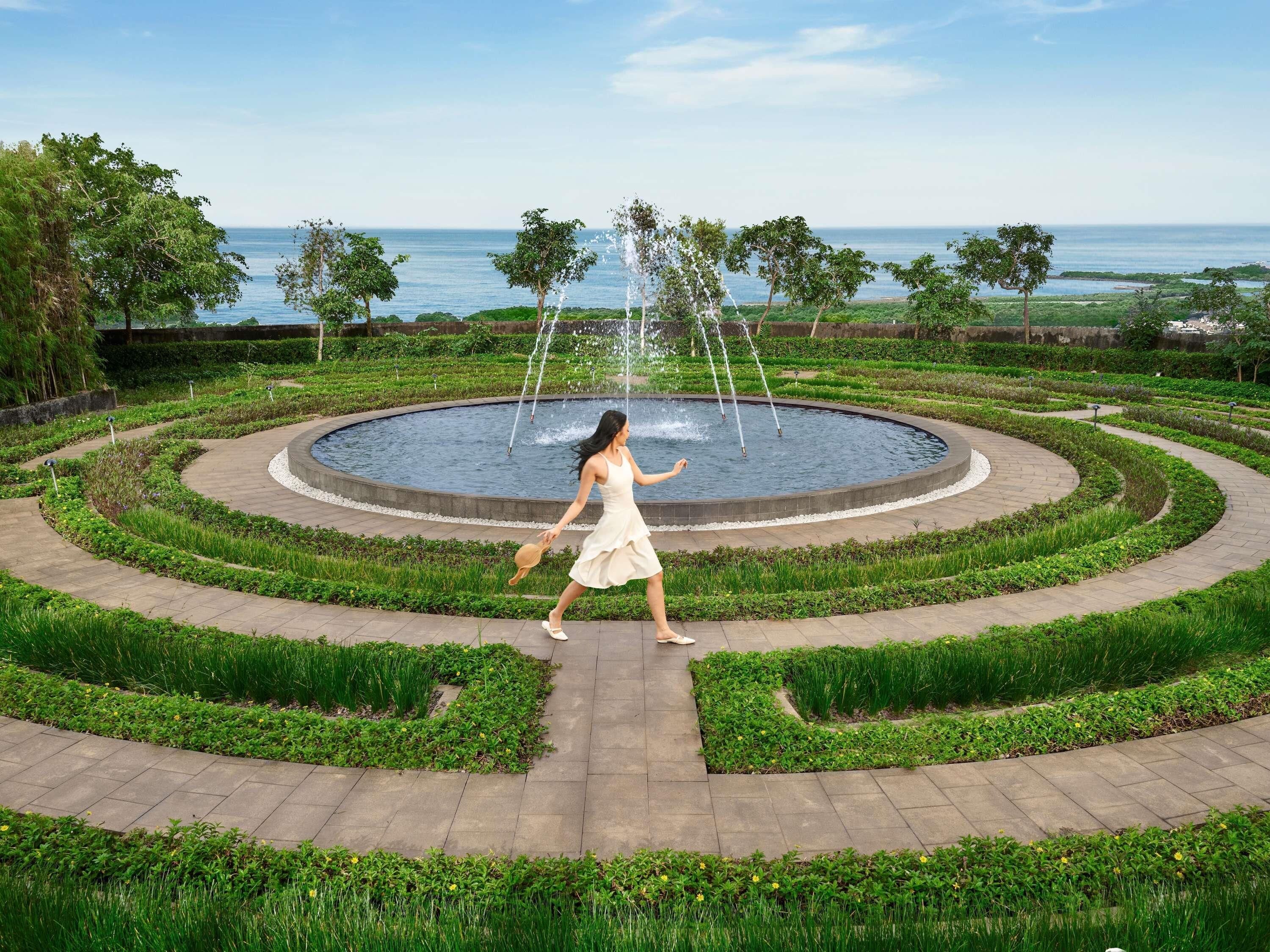 A circular water feature with fountains, surrounded by a landscaped garden with a paved walkway.