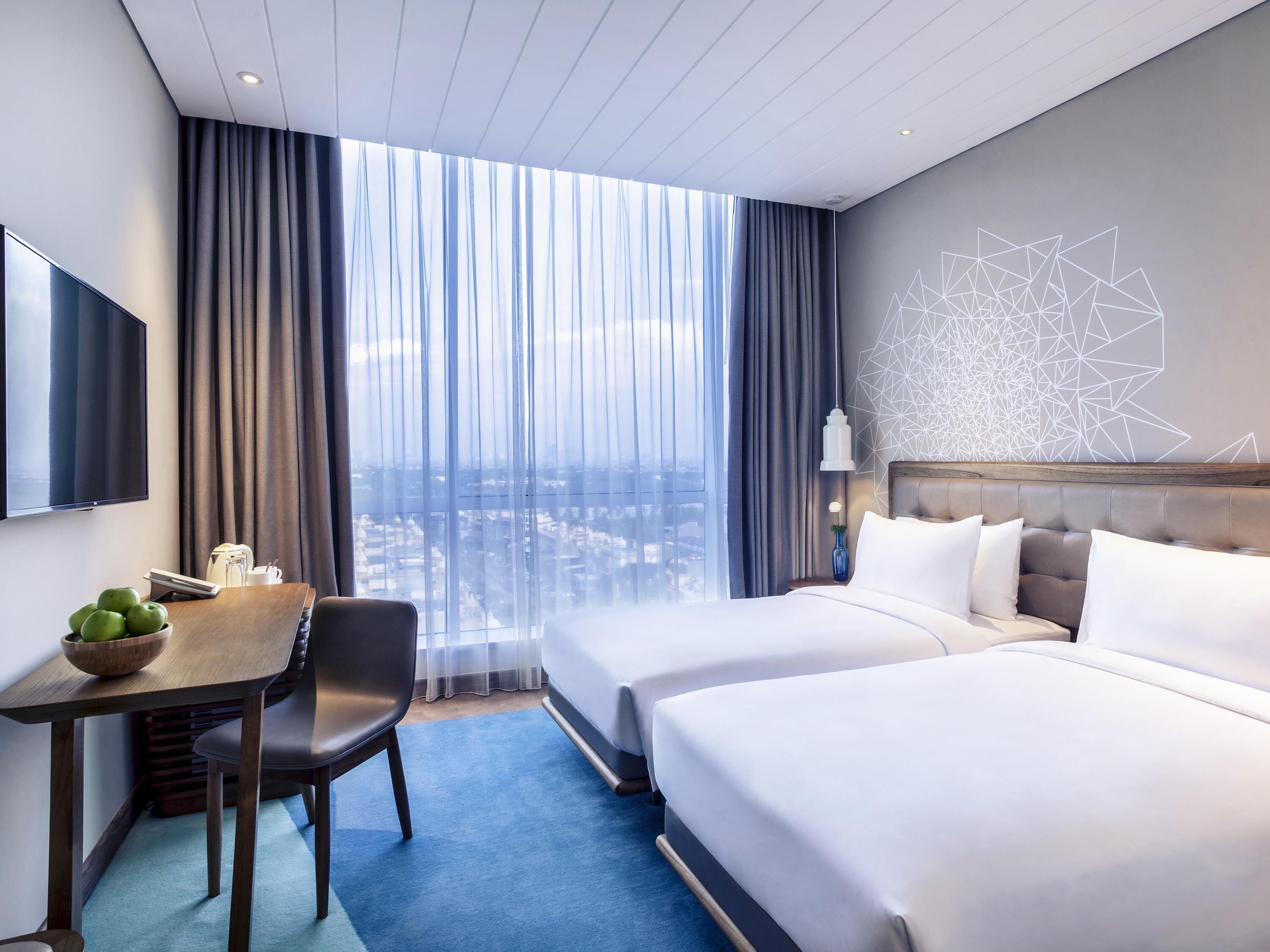 Habitación de hotel con cama, escritorio con frutero, silla, televisión y ventana grande con cortinas.