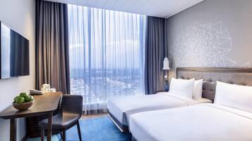 Habitación de hotel con cama, escritorio con frutero, silla, televisión y ventana grande con cortinas.