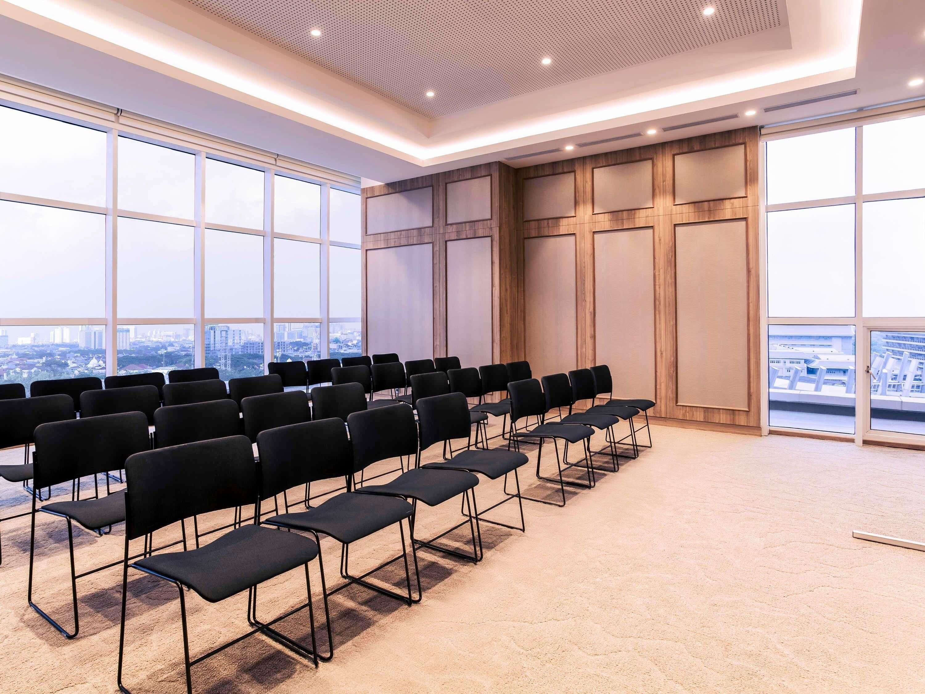 Un salón de conferencias con filas de sillas negras, ventanas amplias y vista a la ciudad.