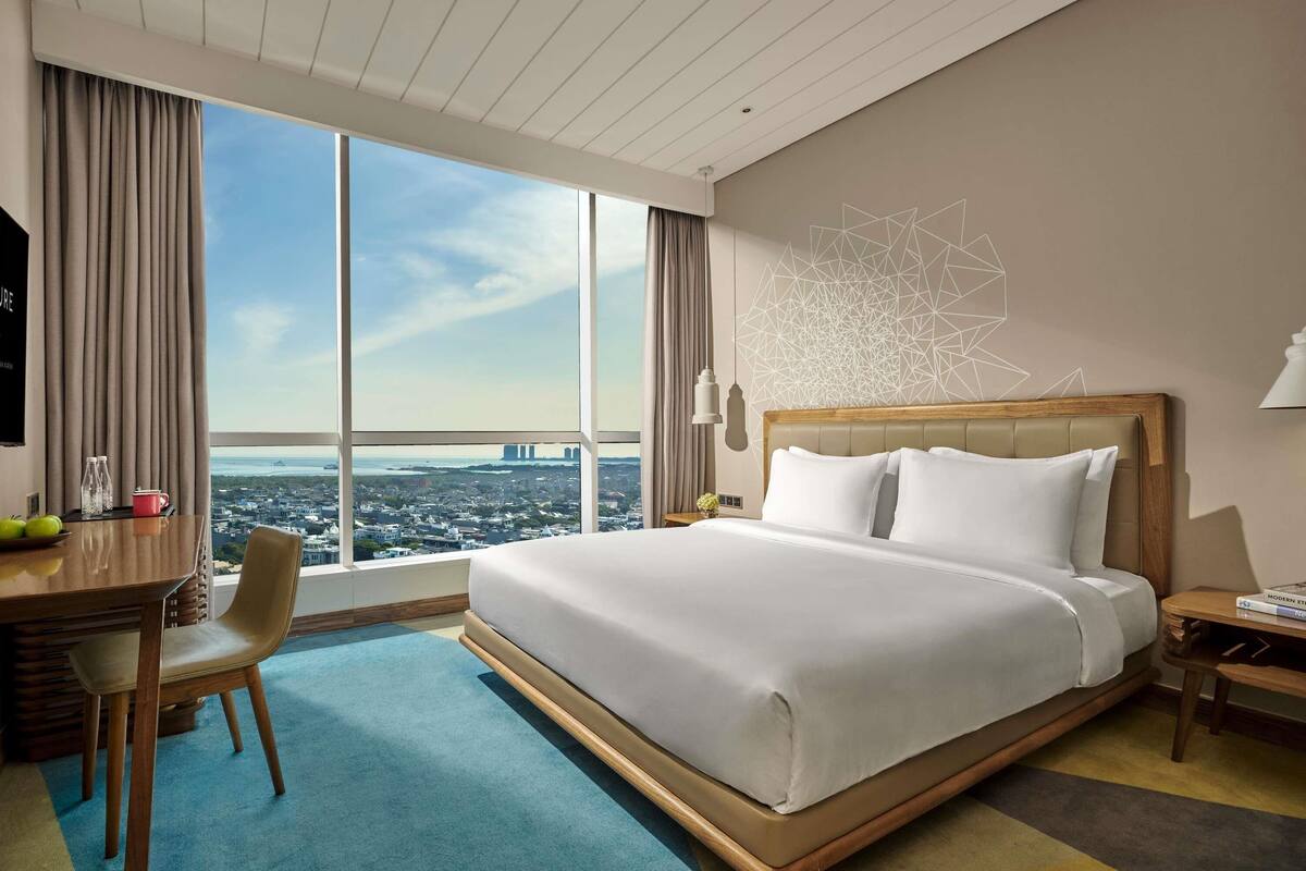 Une chambre d’hôtel avec un grand lit, un bureau avec une chaise, une vue sur le paysage urbain et un motif géométrique sur le mur.