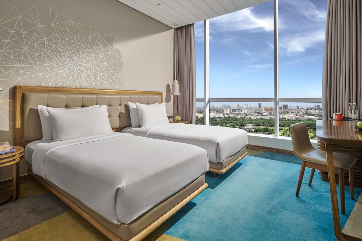Une chambre d’hôtel avec deux lits, une table à manger, une chaise, une vue sur la ville et un motif géométrique sur le mur.