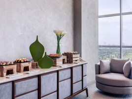 A modern interior with a wooden console table displaying food items, a green vase, and a tall flower arrangement. A grey upholstered armchair is positioned near a large window offering a city view.