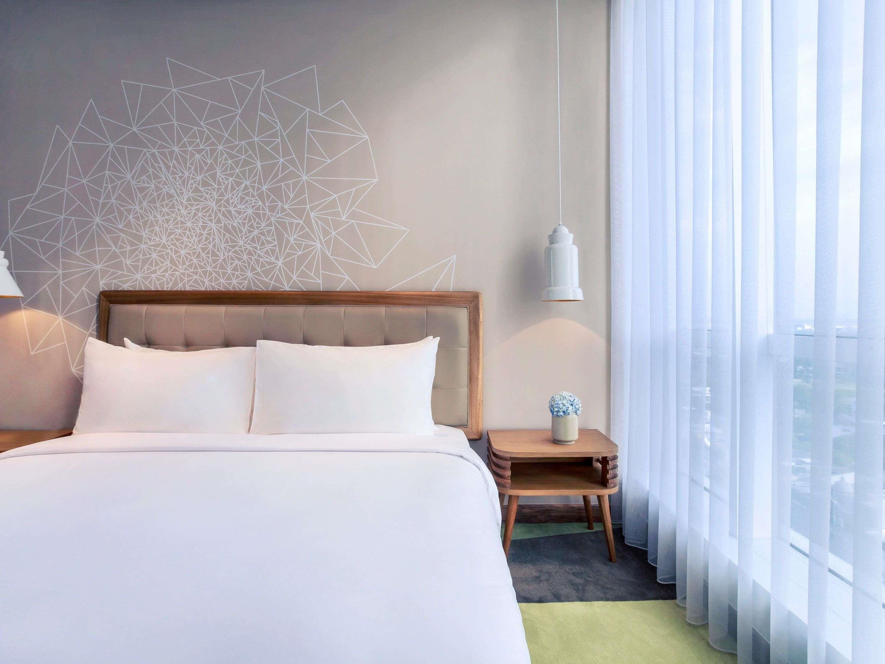 A hotel room with a large bed, a geometric wall design, a bedside table with a lamp, and a view through sheer curtains.