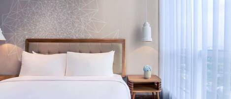 A hotel room with a large bed, a geometric wall design, a bedside table with a lamp, and a view through sheer curtains.