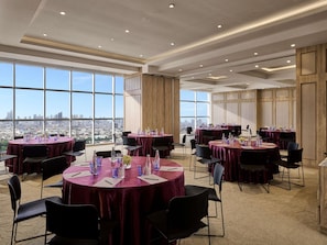 Un salón de conferencias con mesas redondas, sillas y vistas a la ciudad a través de amplios ventanales.
