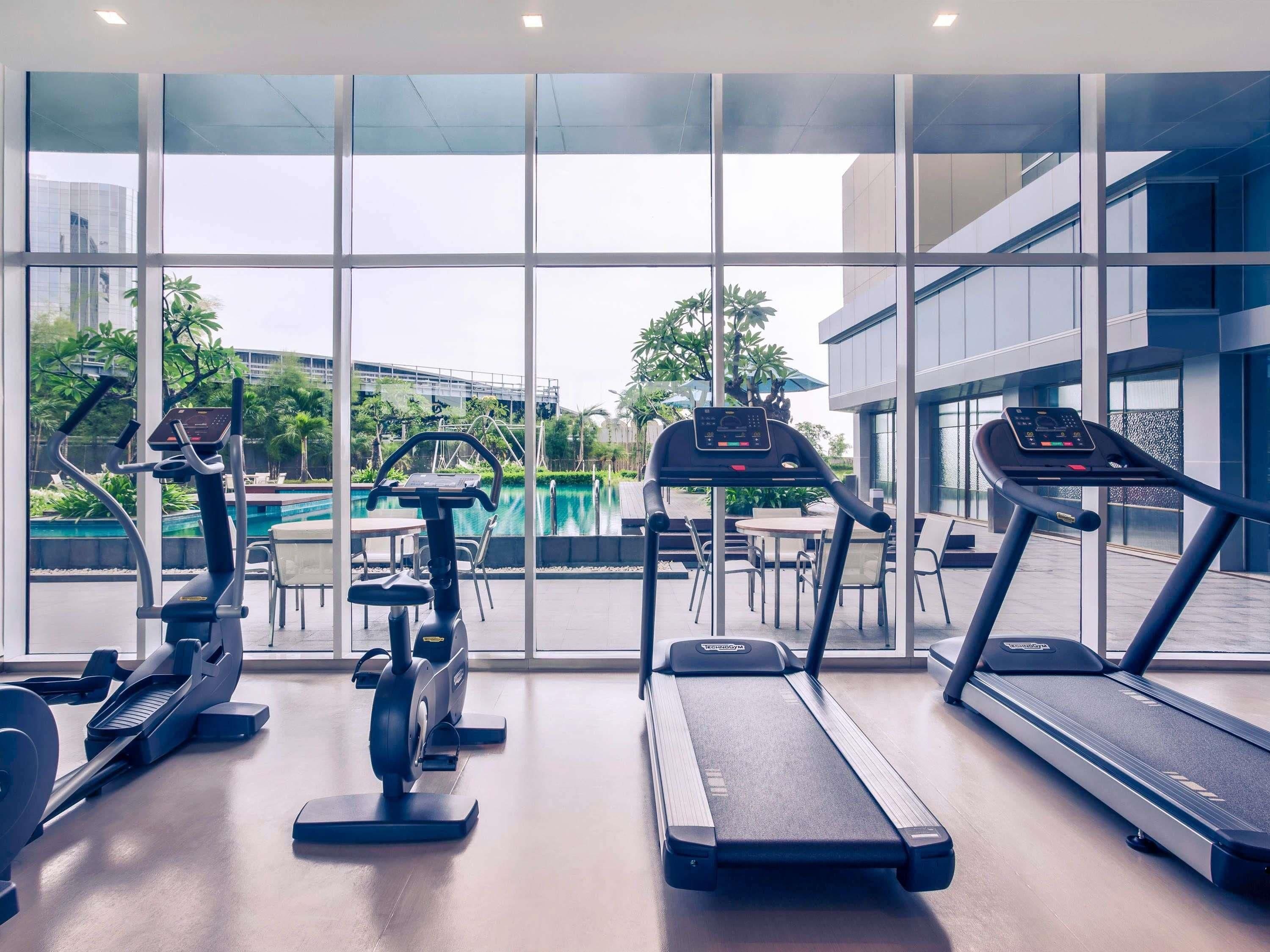 Un gimnasio con cintas de correr, elíptica y remo, con vista a una piscina y palmeras.