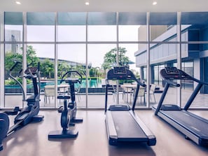 Une salle de sport équipée de tapis de course, d’un vélo elliptique et d’un rameur, avec vue sur une piscine et des palmiers.