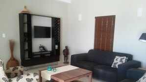 Una sala de estar con un sofá de color oscuro, una mesa de centro de madera, un sillón estampado y un televisor sobre un soporte negro.
