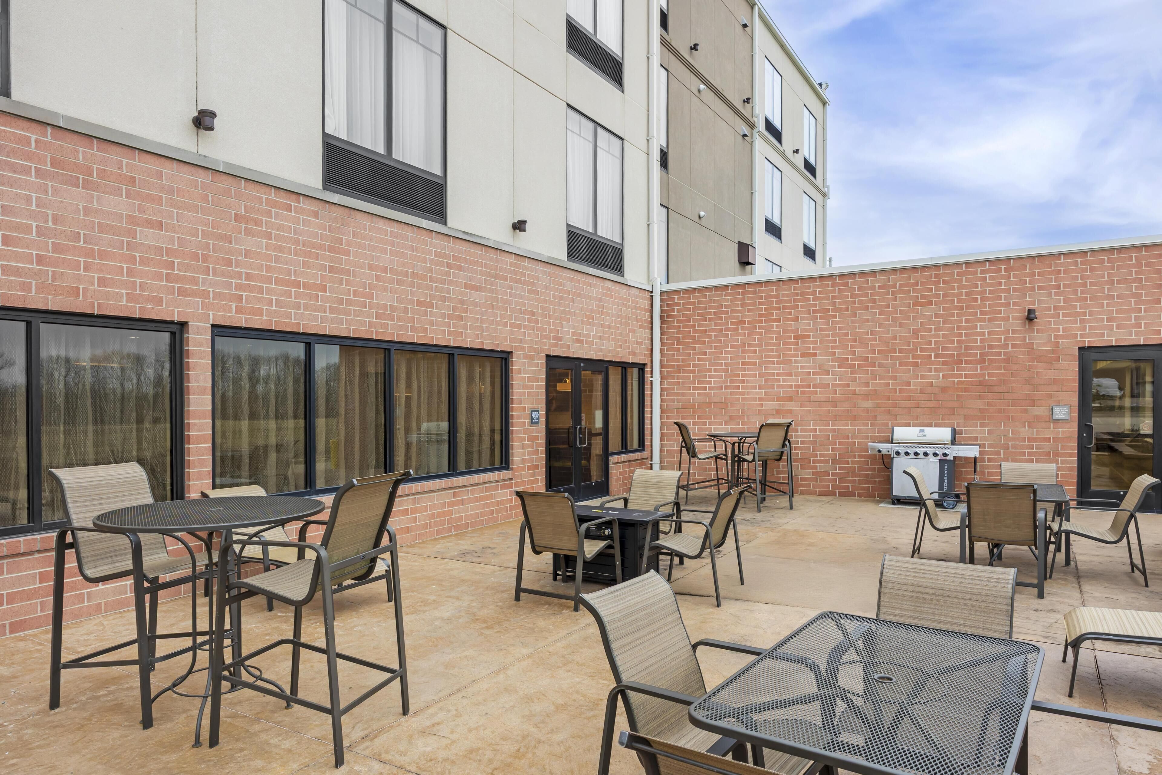 An outdoor patio with tables and chairs, a grill, and a brick building in the background.