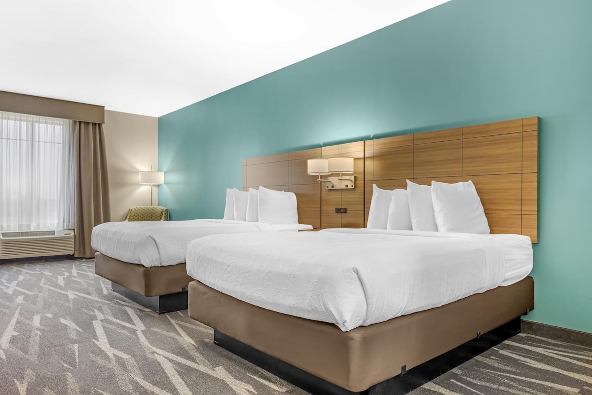 A hotel room with two beds, a large window with curtains, a wall-mounted lamp, and a patterned carpet.