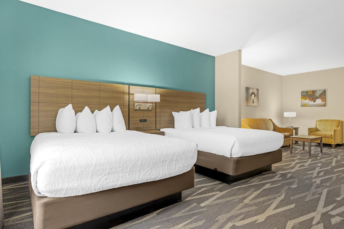 A hotel room with two beds, a wooden headboard, a teal accent wall, a patterned carpet, and a small seating area with a chair and a table.