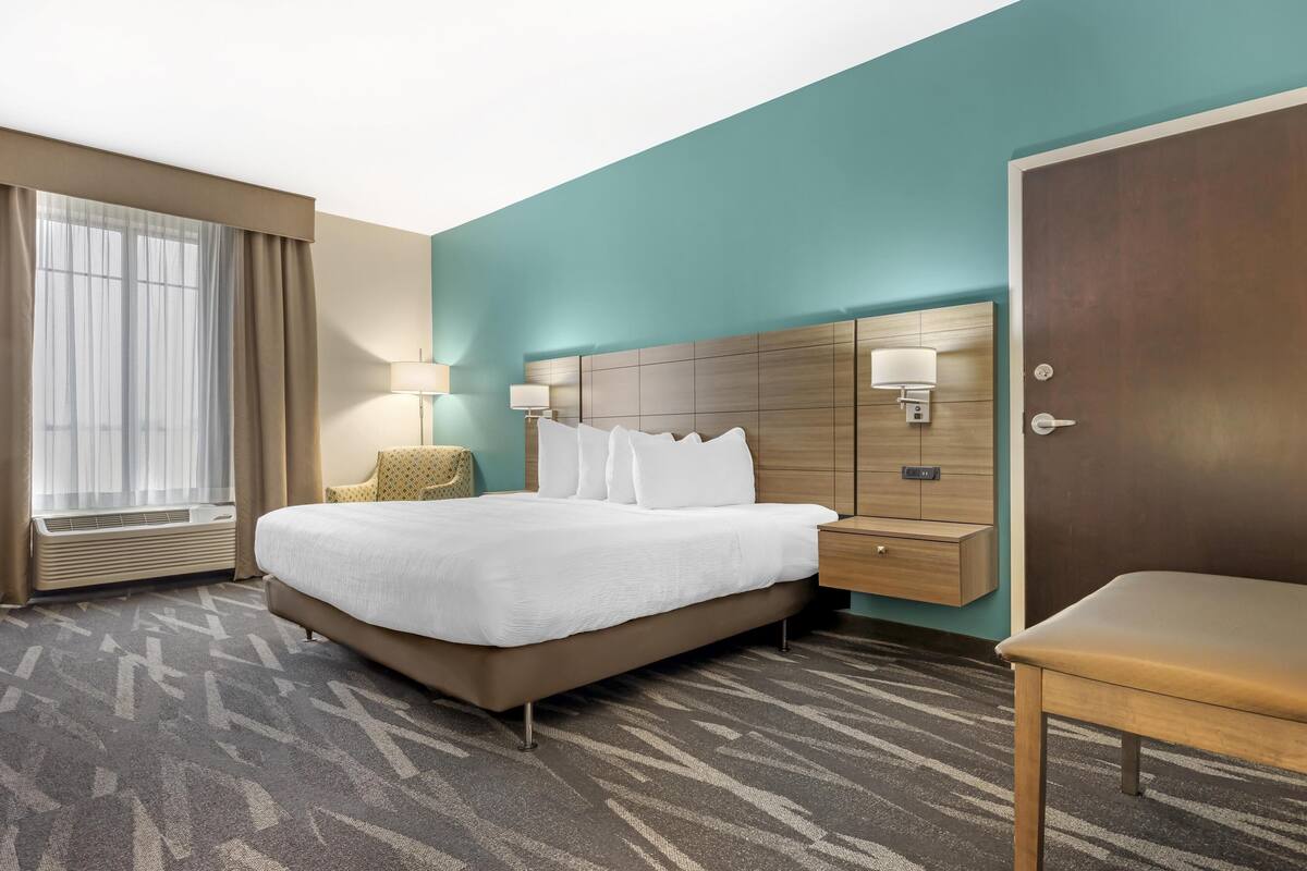A hotel room with a large bed, a wooden nightstand, a bedside table, a chair, and a door.