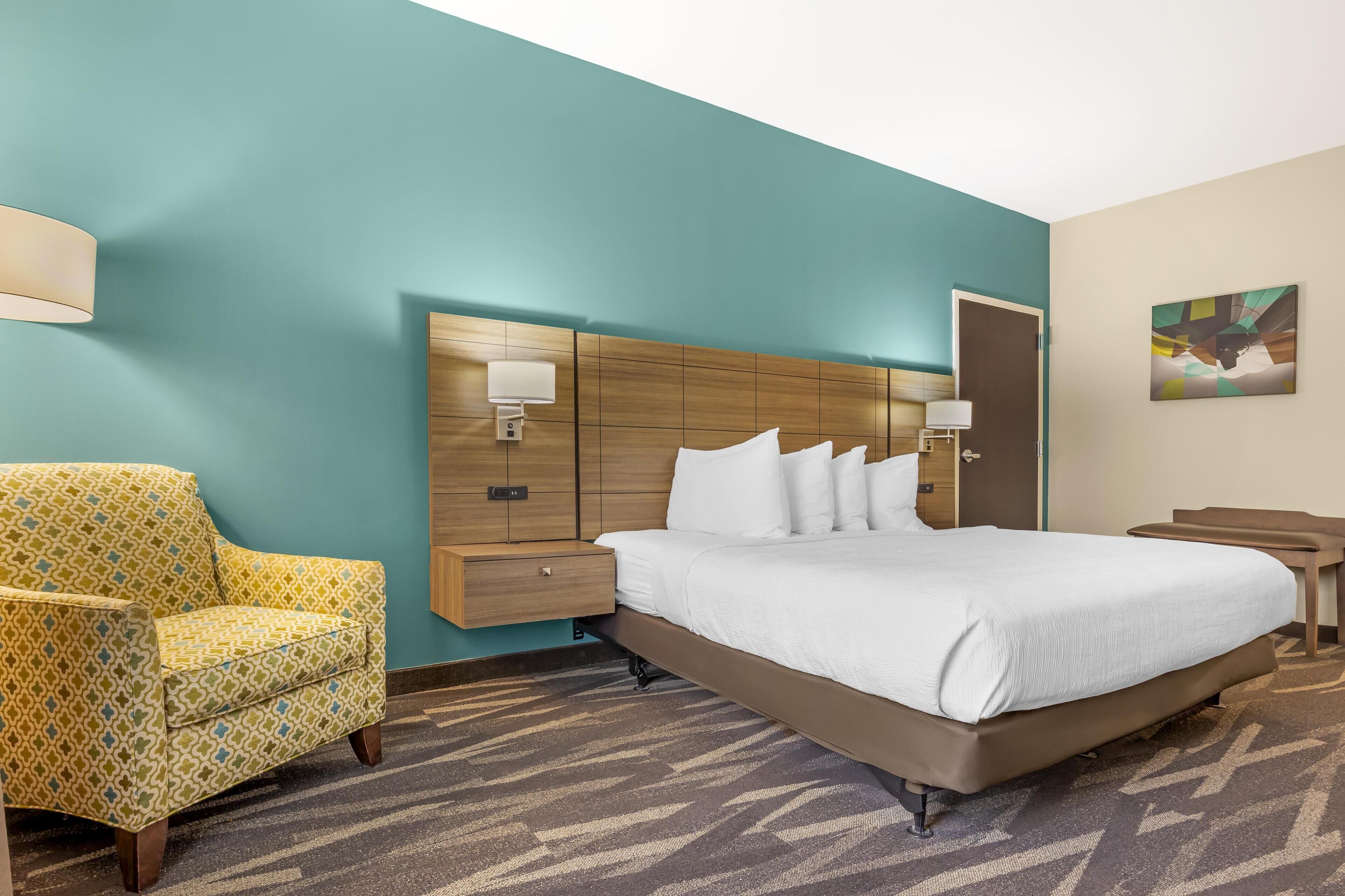 A hotel room with a large bed, a patterned armchair, a wooden headboard, and a wall-mounted lamp.