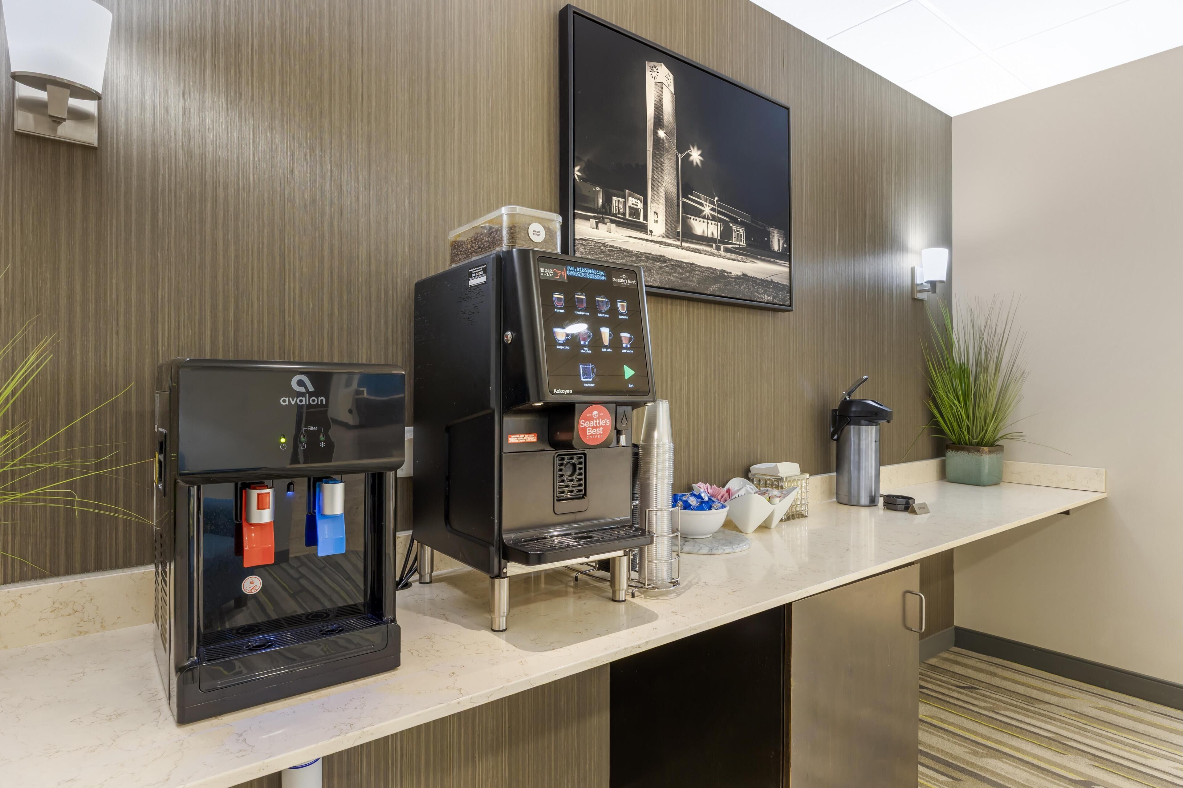 A hotel reception area with a coffee machine, a beverage dispenser, and a framed picture on the wall.