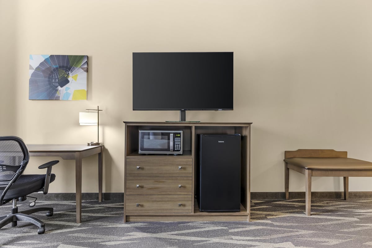 A hotel room with a flat-screen TV mounted on the wall, a microwave, and a refrigerator on a wooden cabinet, a desk with a chair, and a patterned carpet.