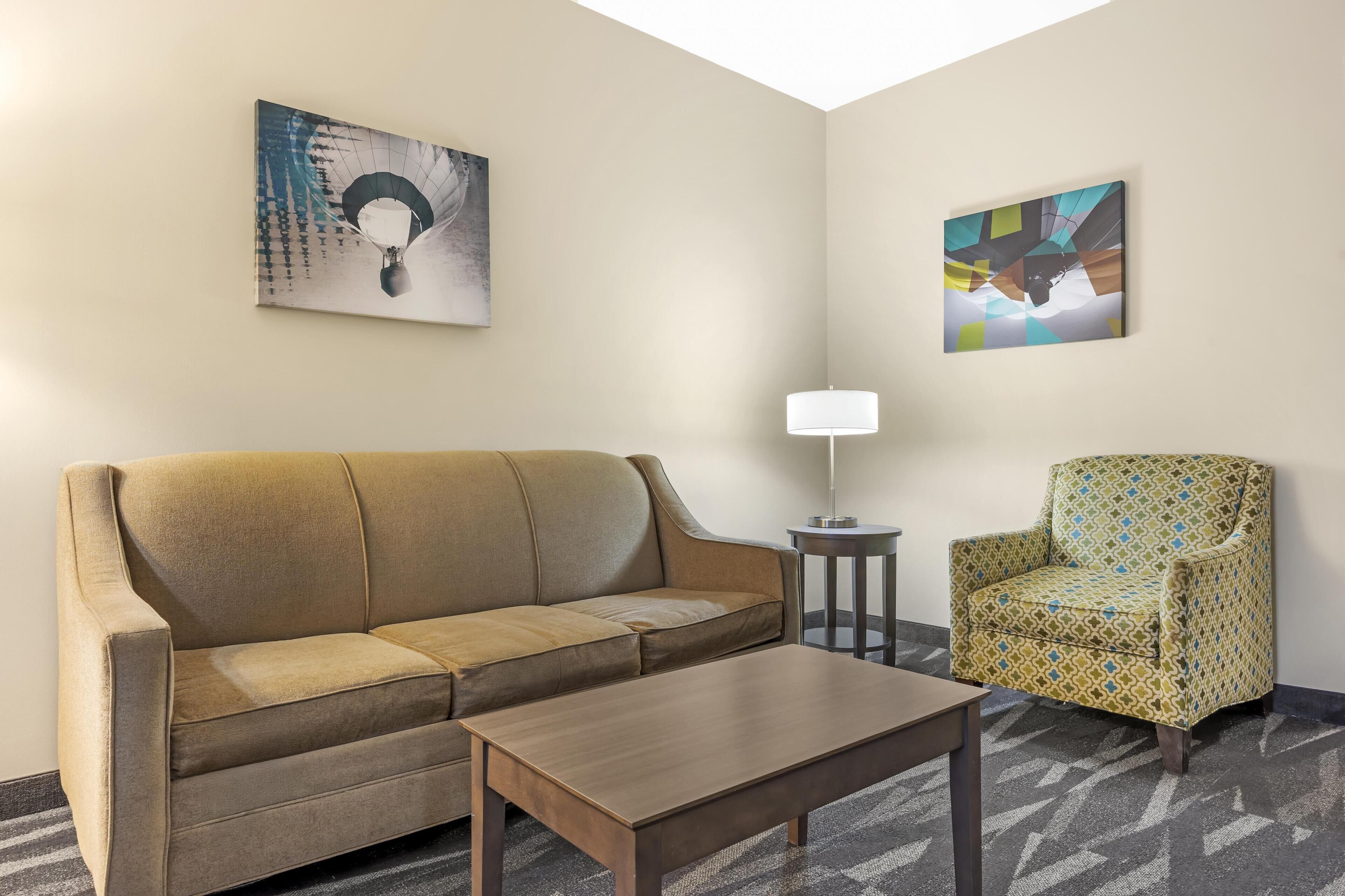 Habitación de hotel con sofá color beige, mesa de centro de madera, sillón estampado, lámpara de pie y dos cuadros abstractos en las paredes.