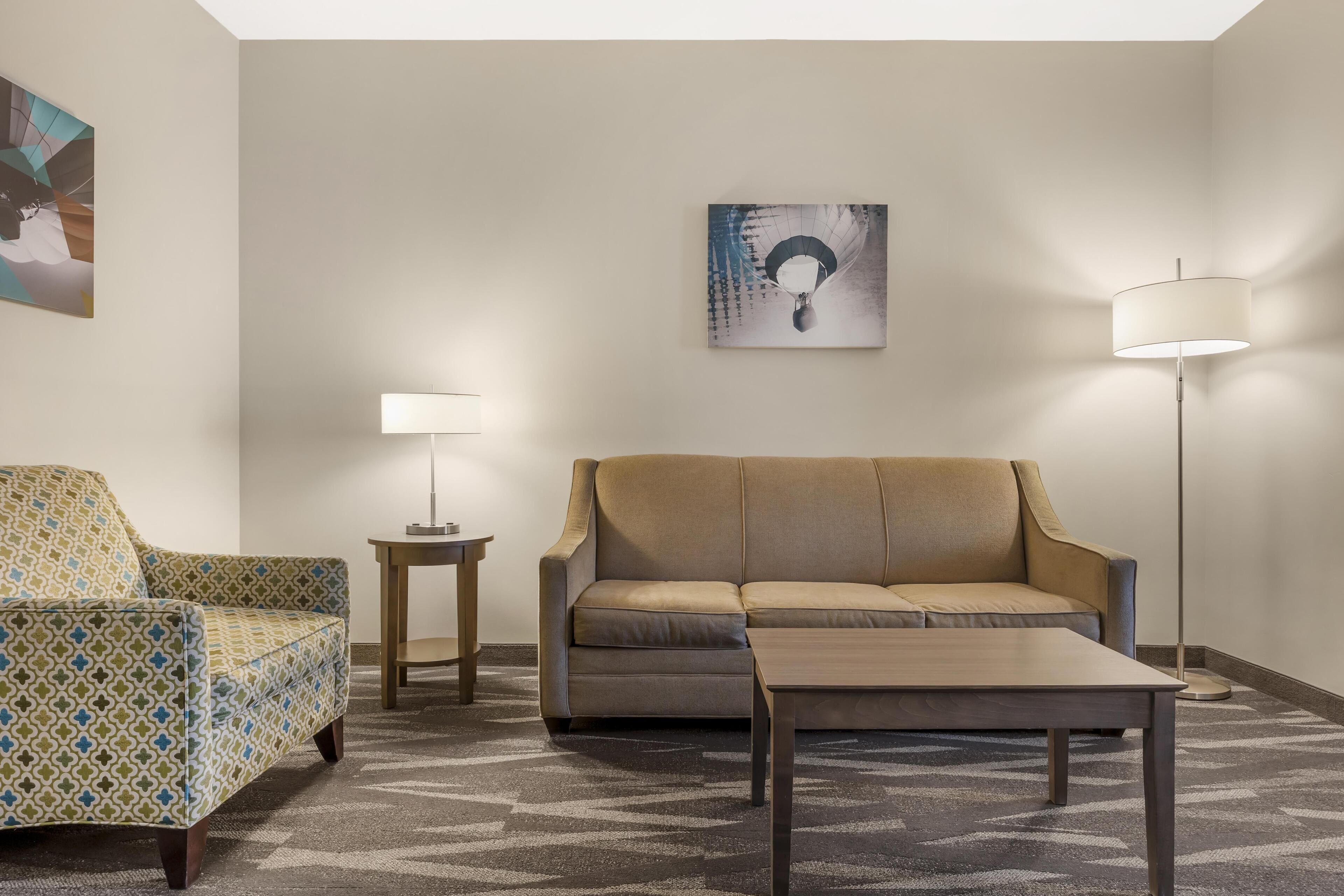 A hotel room with a patterned armchair, a sofa, a coffee table, a floor lamp, and a painting on the wall.