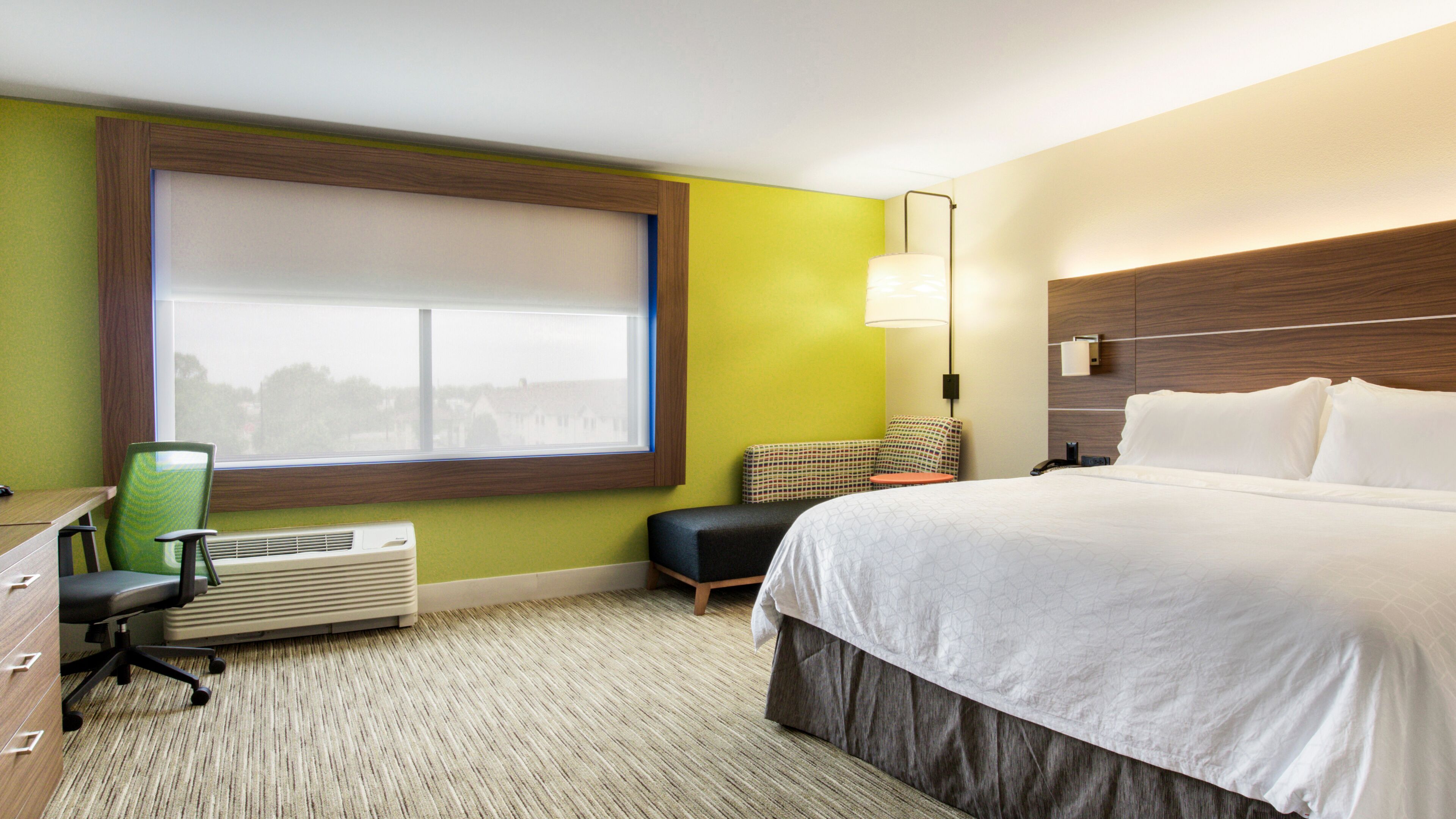 A hotel room with a large bed, a desk with a chair, a small table, a lamp, and a window with blinds.