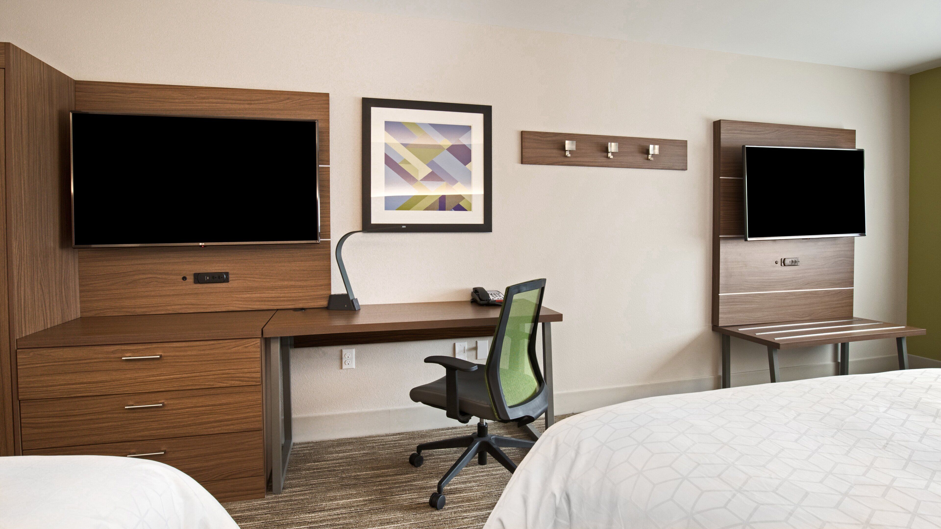 Ein Hotelzimmer mit einem Holztisch, zwei Betten, einem Sessel, einem Flachbildfernseher, einem gerahmten Bild und einem Telefon.