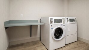 A laundry room with two front-loading washing machines, a metal shelf, and a wall socket. - Holiday Inn Express & Suites Racine by IHG (Mount Pleasant)