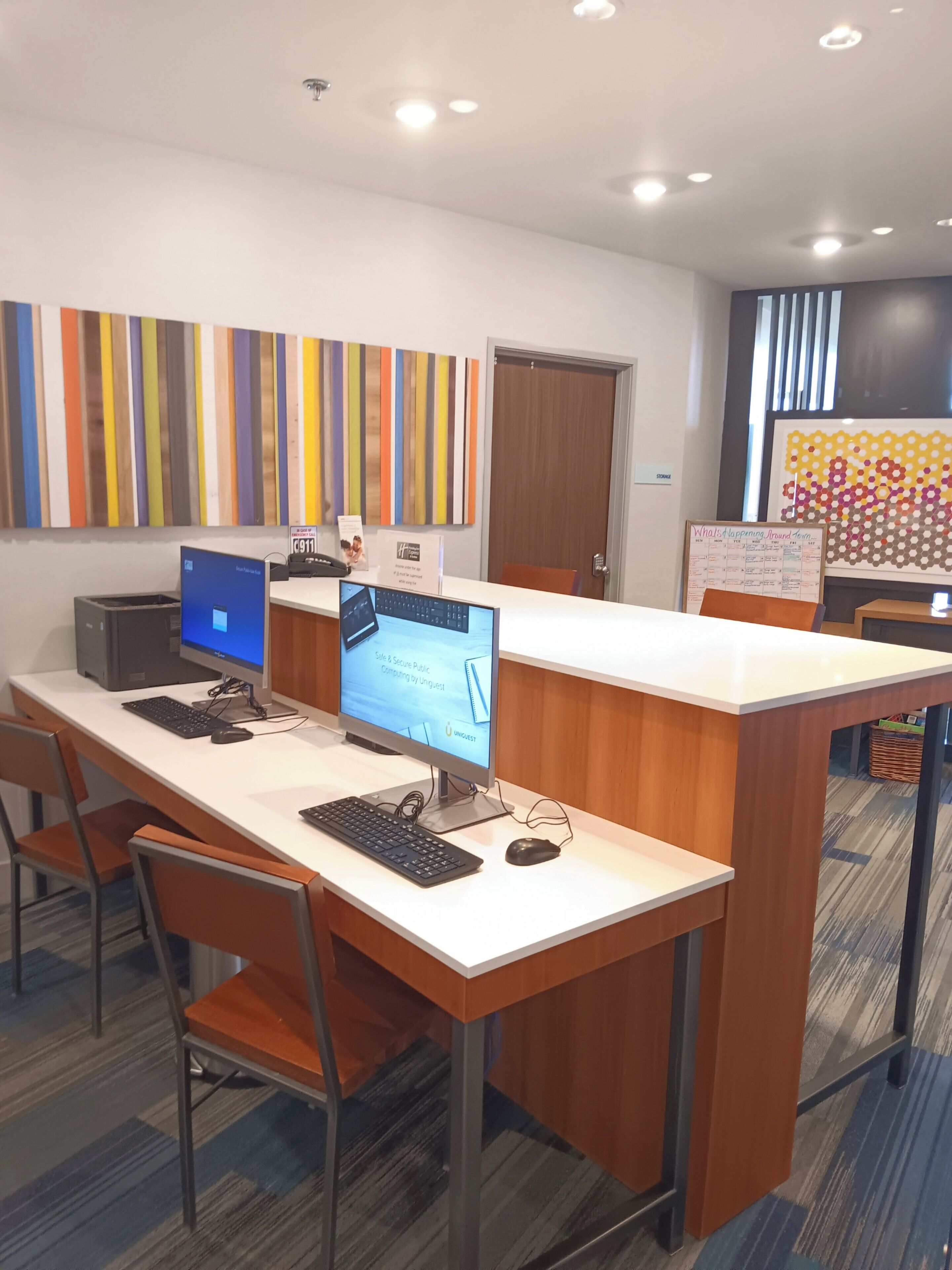 Un espai d'oficina modern amb dues taules, cadascuna equipada amb un ordinador i un teclat. Hi ha dues cadires, una amb coixí taronja. La sala destaca per un quadre de paret amb dibuixos de tires de colors, una porta de fusta i un tabló d'anuncis amb un calendari.