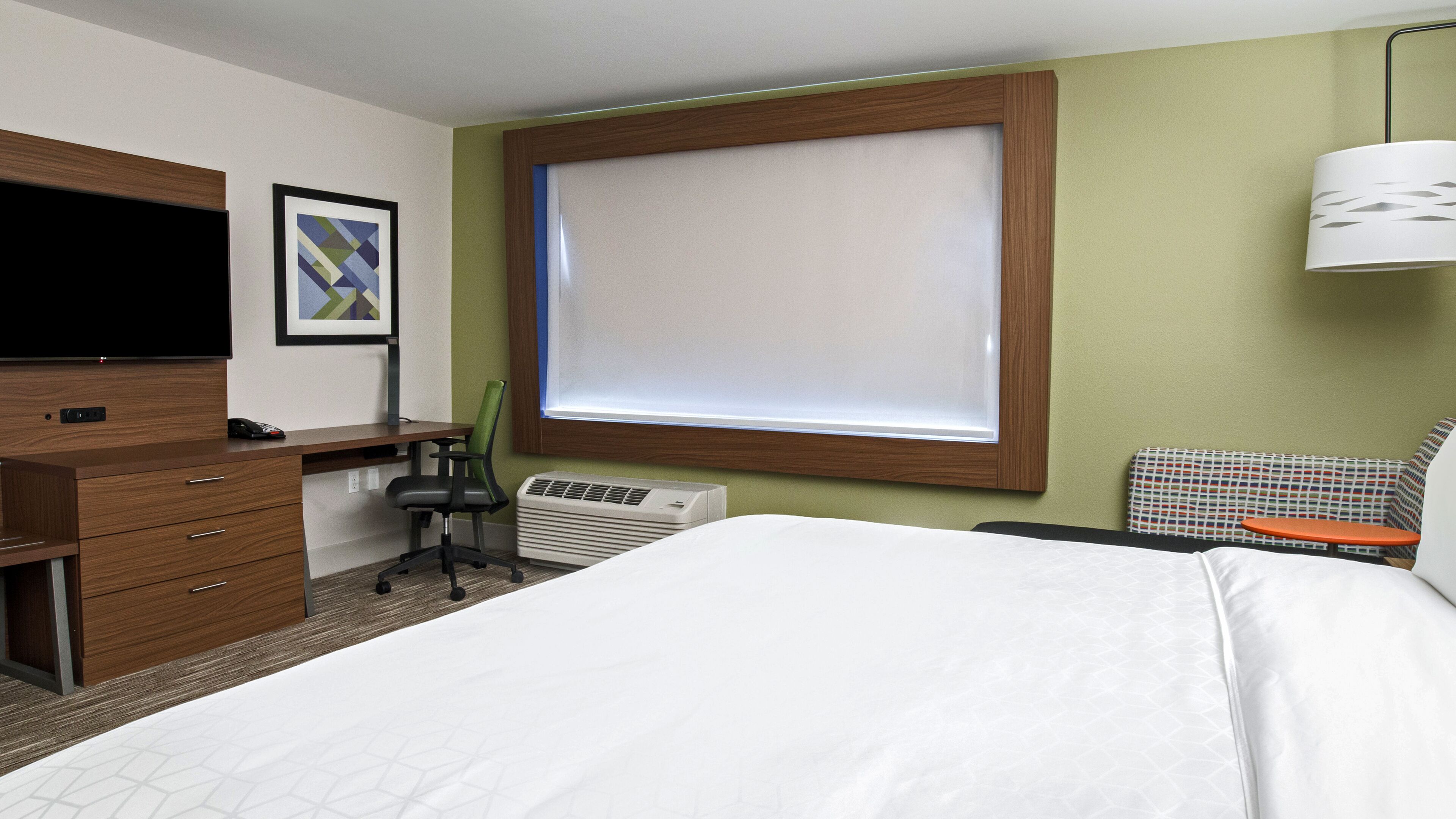 A hotel room with a bed, desk, chair, TV, air conditioning unit, and a window with blinds.