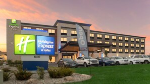 A Holiday Inn Express & Suites building with a sign displaying the logo and name, a parking lot with several cars, and a flagpole with a flag.
