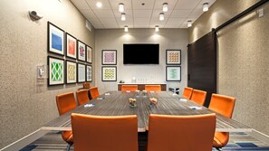 Ett konferensrum med ett långt bord, orange stolar, inramade konstverk på väggarna och en platt-TV.