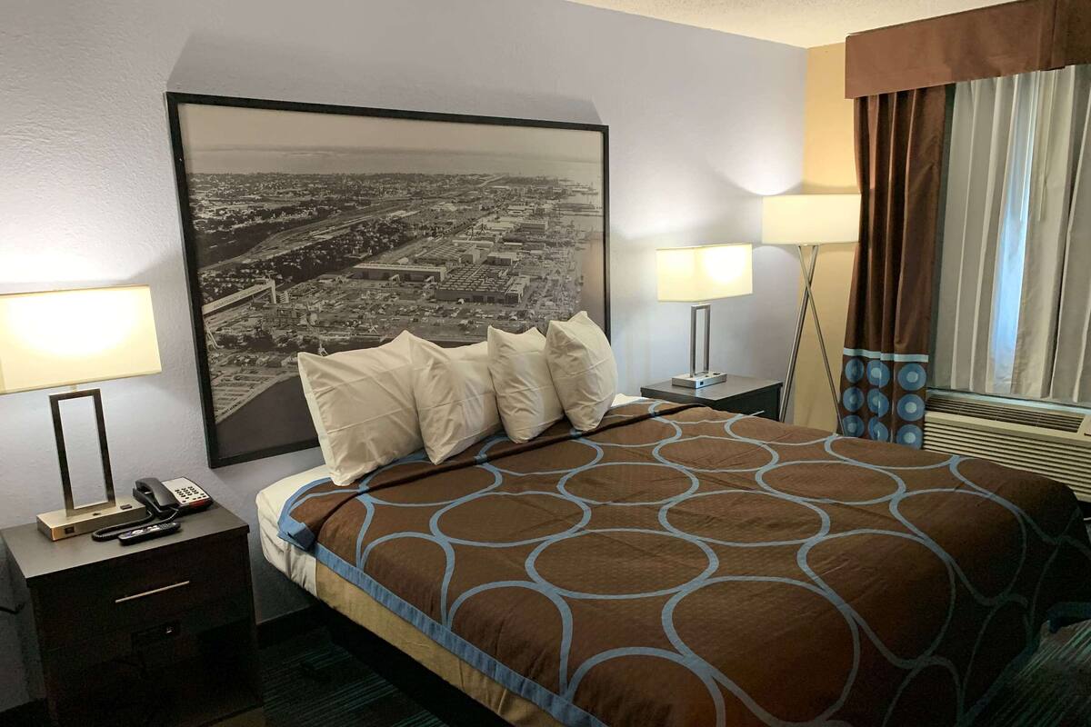 Una habitación de hotel con una cama grande, una mesita de noche con un teléfono y controles remotos, una lámpara de escritorio y un cuadro grande con una vista urbana enmarcada en la pared.