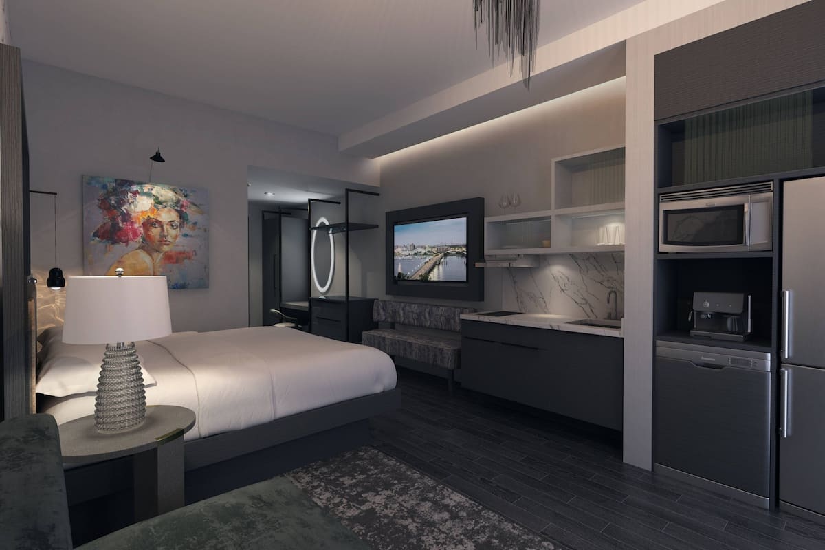 A modern bedroom with a large bed, a TV mounted on the wall, a built-in kitchen with a microwave, and a bathroom with a mirror and shower.