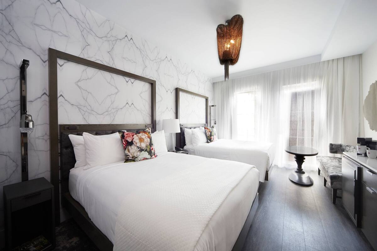 A hotel room with two beds, a dining table, a small kitchen area, and a large window with curtains.