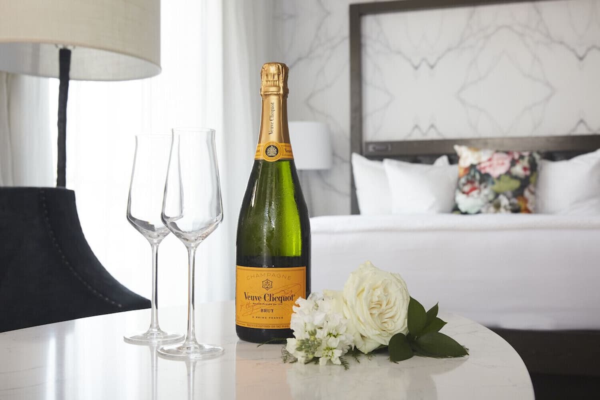 A bottle of Veuve Clicquot champagne, two empty glasses, and a white rose on a table in a hotel room.