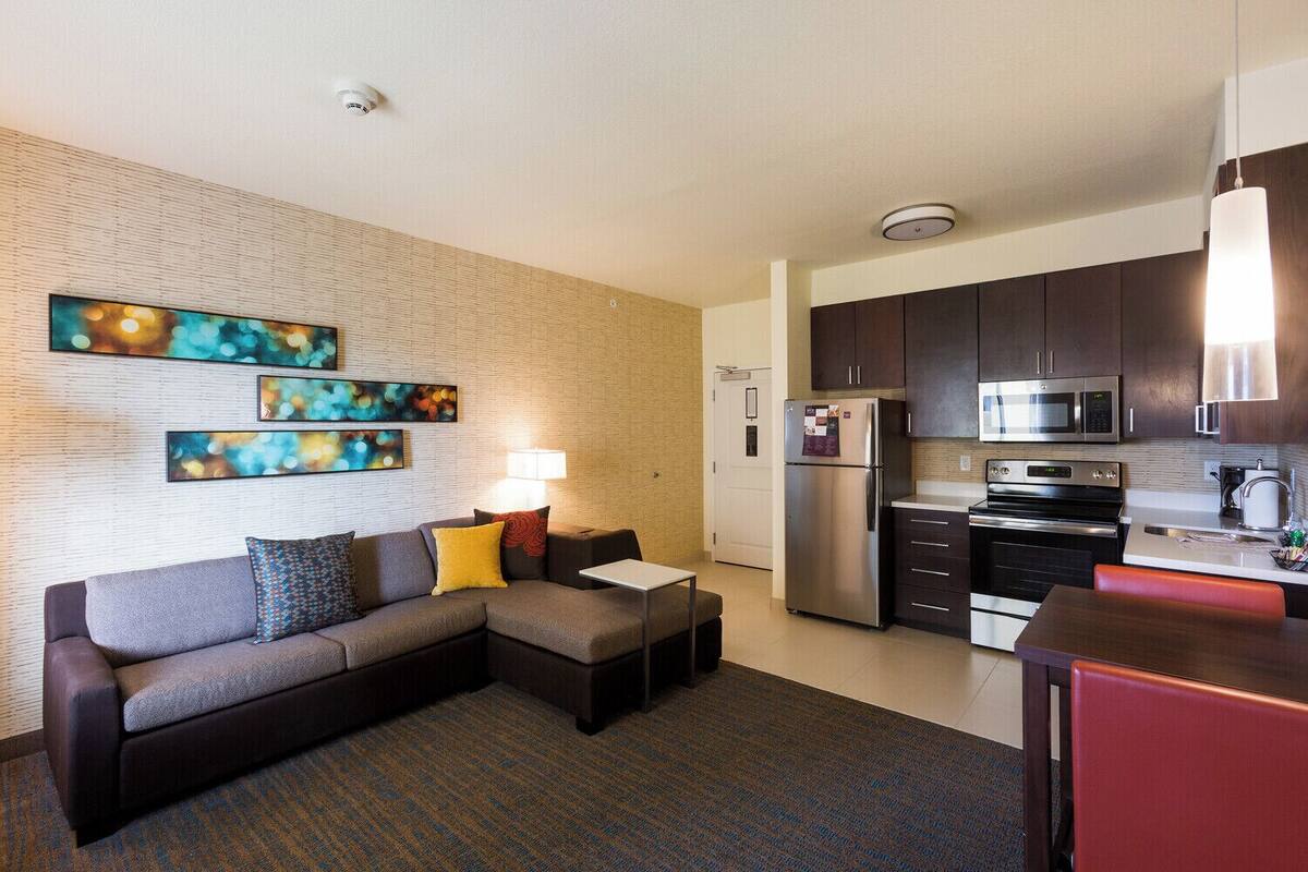 A modern living room with a sectional sofa, a small table, and a lamp. Adjacent is a compact kitchen with a refrigerator, microwave, and stove.